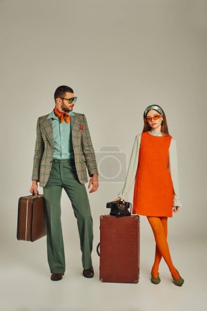old-fashioned couple in bright clothes posing with vintage suitcases and retro telephone on grey