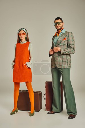 Frau in orangefarbenem Kleid und Sonnenbrille neben glücklichem Mann im karierten Blazer und Vintage-Koffer auf grau