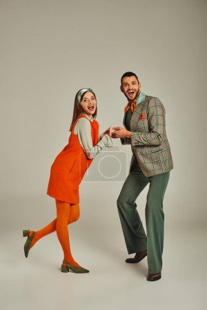 full length of overjoyed couple in elegant retro attire looking at camera and holding hands on grey