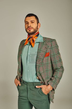 confident man in plaid blazer and colorful neckerchief standing with hands in pockets on grey
