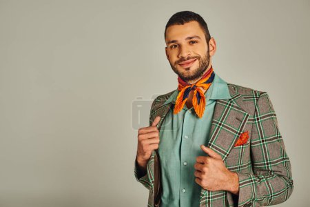 elegant man in checkered blazer and colorful neckerchief smiling at camera on grey, vintage fashion