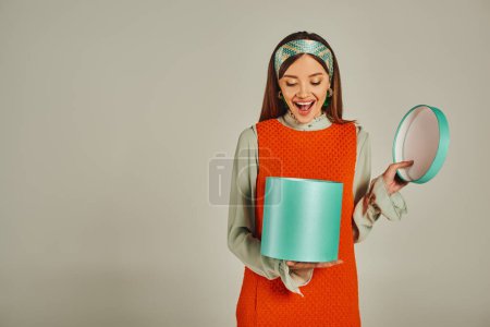 amazed woman in orange dress and colorful headband opening gift box on grey, retro-inspired fashion
