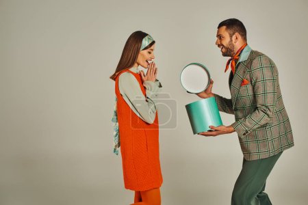 Téléchargez les photos : Homme tendance ouverture boîte cadeau près surprise femme en robe orange sur gris, couple d'inspiration rétro - en image libre de droit