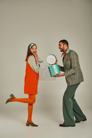 Photo for Stylish man holding gift box near overjoyed woman in orange dress on grey, retro-inspired fashion - Royalty Free Image