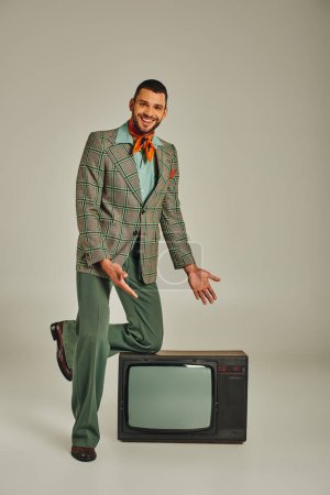 smiling man in elegant retro style attire pointing at vintage tv set on grey backdrop, full length
