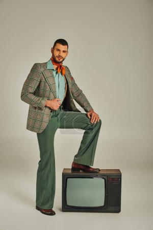 hombre seguro de sí mismo en ropa retro con estilo pisando el televisor vintage y mirando a la cámara en gris