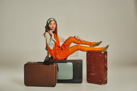 mujer de estilo retro sentado en maletas vintage y televisor mientras habla en el teléfono con cable en gris