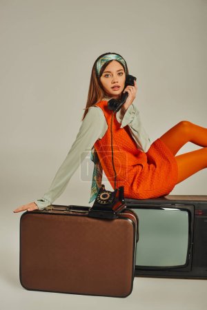 charming retro woman sitting on vintage suitcase and tv set while talking on telephone on grey