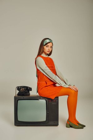 trendy woman in retro style clothes sitting on vintage tv set near corded phone on grey, full length