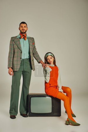 confident man holding hands with charming woman sitting on vintage tv set on grey, retro lifestyle