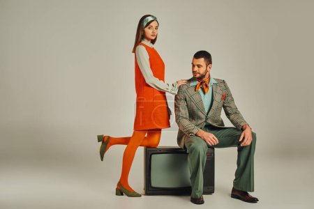 Frau in orangefarbenem Kleid berührt Schulter des Mannes sitzend auf Vintage-TV-Set auf grau, Retro-Stil