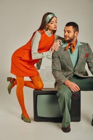 charming woman leaning on shoulder of man sitting on vintage tv on grey, trendy retro couple