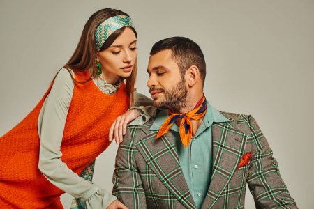 woman in orange dress leaning on shoulder of man in plaid jacket on grey, stylish vintage couple