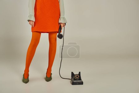 Ausgeschnittene Ansicht einer Frau in orangefarbenem Kleid und Strumpfhose mit Hörer eines alten Telefons auf grau, Retro-Stil
