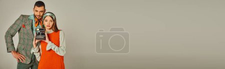 Téléchargez les photos : Élégant jeune couple en tenue rétro avec caméra vintage regardant caméra sur gris, bannière - en image libre de droit