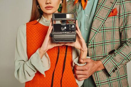 cropped view of woman in orange dress holding vintage camera near man in plaid jacket on grey