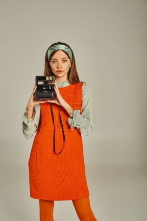 trendy woman in orange and colorful headband holding vintage camera on grey, retro-inspired style