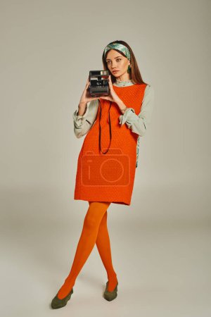 full length of woman in bright retro clothes taking photo on vintage camera on grey backdrop