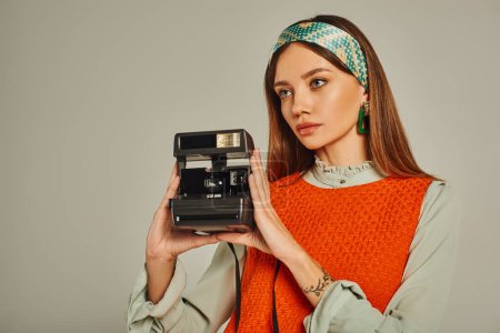 bezaubernde Frau in buntem Stirnband und orangefarbenem Kleid beim Fotografieren auf Vintage-Kamera auf grau
