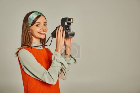 fröhliche Frau in orangefarbenem Kleid und hellem Stirnband beim Fototermin mit Vintage-Kamera auf grau im Retro-Stil