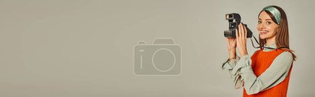 heureuse femme de style rétro en robe orange et bandeau prenant des photos sur appareil photo vintage sur gris, bannière