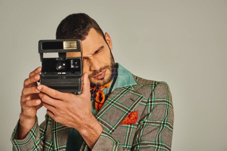 man in plaid blazer and colorful neckerchief taking photo on vintage camera on grey, retro fashion