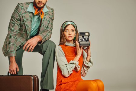 femme en robe orange tenant caméra vintage près de l'homme avec valise sur gris, mode rétro