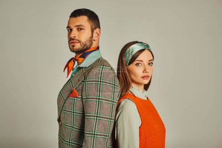 junges Paar in stylischer altmodischer Kleidung, Rücken an Rücken stehend und in die Kamera auf grau blickend