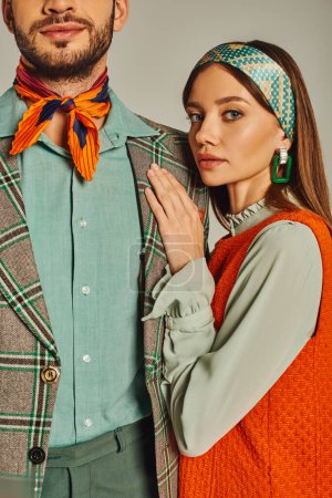 young woman in orange dress looking at camera near man in plaid jacket on grey, fashion from past
