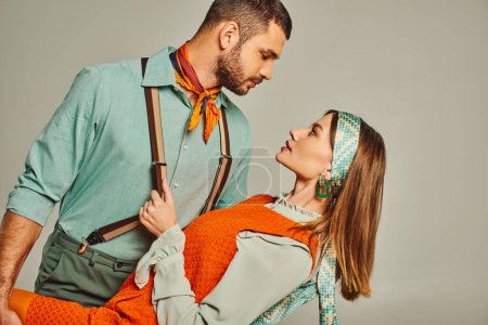 Téléchargez les photos : Vue latérale de style rétro homme en bretelles flirter avec femme en robe orange sur gris, couple de hanche - en image libre de droit