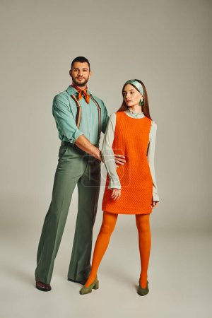 Vintage-Mode, volle Länge des Mannes in Hosenträgern umarmt Taille der Frau in orangefarbenem Kleid auf grau