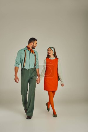 romantisches Paar in eleganter Retro-Kleidung, Händchen haltend, spazierend und einander lächelnd auf grau