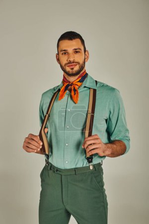 hombre positivo en tirantes y colorido pañuelo mirando a la cámara en gris, estilo retro-inspirado