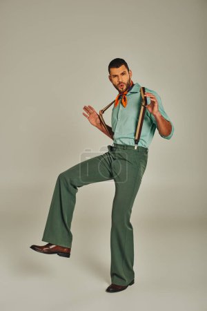 excited hip man in stylish vintage clothes pulling suspenders and looking at camera on grey