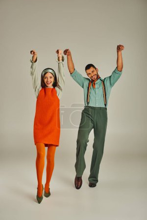 cheerful couple in vintage clothes dancing with raised hands on grey, happiness and retro vibes