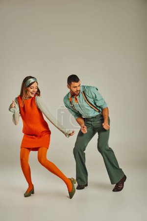 full length of young fashionable couple in colorful retro attire dancing on grey, fun and excitement