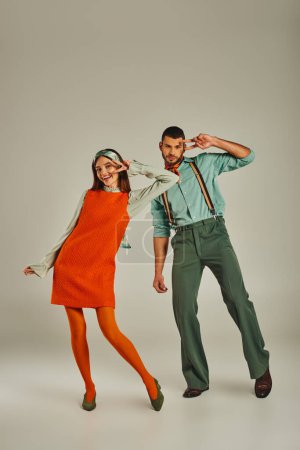 aufgeregtes junges Paar im Vintage-Outfit zeigt Siegeszeichen und tanzt auf grauem Retro-Lifestyle