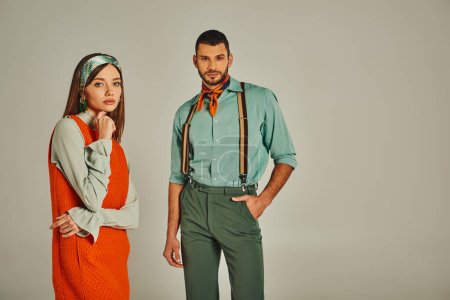 confident man with hand in pocket near thoughtful woman in orange dress on grey, retro fashion