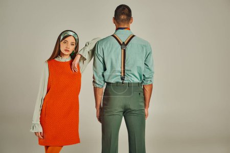 junge Frau in orangefarbenem Kleid blickt in die Kamera neben trendigen Mann in Hosenträgern auf grau, Vintage-Stil