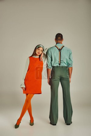 woman in orange dress looking at camera near stylish man in suspenders on grey, old-fashioned couple