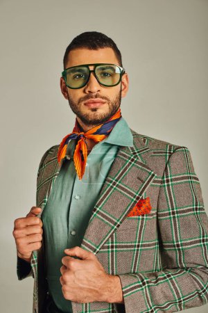 Mann im Retro-Stil in kariertem Blazer und buntem Halstuch mit Sonnenbrille blickt in die Kamera auf grau