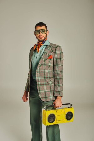 Téléchargez les photos : Homme à la mode en blazer à carreaux et lunettes de soleil tenant boombox jaune sur gris, style de vie rétro-inspiré - en image libre de droit
