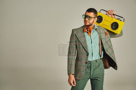 eleganter Musikliebhaber in karierter Jacke und Sonnenbrille mit gelber Boombox auf grau im Retro-Stil