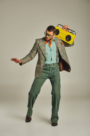 full length of young and trendy old-fashioned man with yellow boombox dancing on grey, retro style