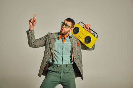 Aufgeregter Mann mit Sonnenbrille, gelbe Boombox in der Hand, nach oben zeigend und auf grau, Vintage-Stil tanzend