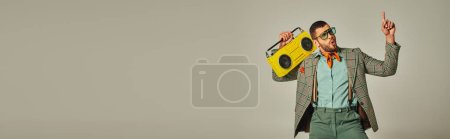excited stylish man in sunglasses holding vintage boombox, pointing up and dancing on grey, banner