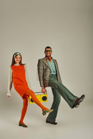 pareja llena de alegría en elegante atuendo retro sosteniendo boombox amarillo y bailando sobre fondo gris