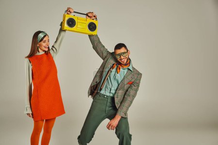 excited couple in stylish vintage clothes holding yellow boombox and dancing on grey backdrop
