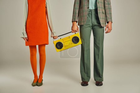 cropped view of couple in colorful clothes standing with yellow boombox on grey, vintage lifestyle