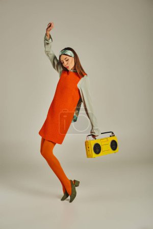 aufgeregte Frau in orangefarbenem Kleid tanzt mit gelber Boombox und erhobener Hand auf grau, Vintage-Vibes